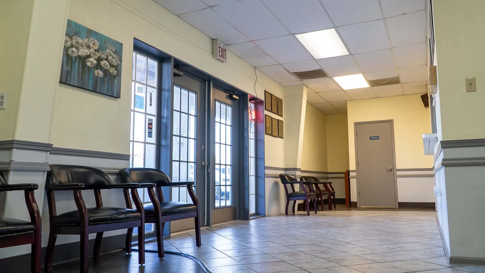 Empty waiting room with black chairs, tiled floor, blue double doors, and wall art under fluorescent ceiling lights.