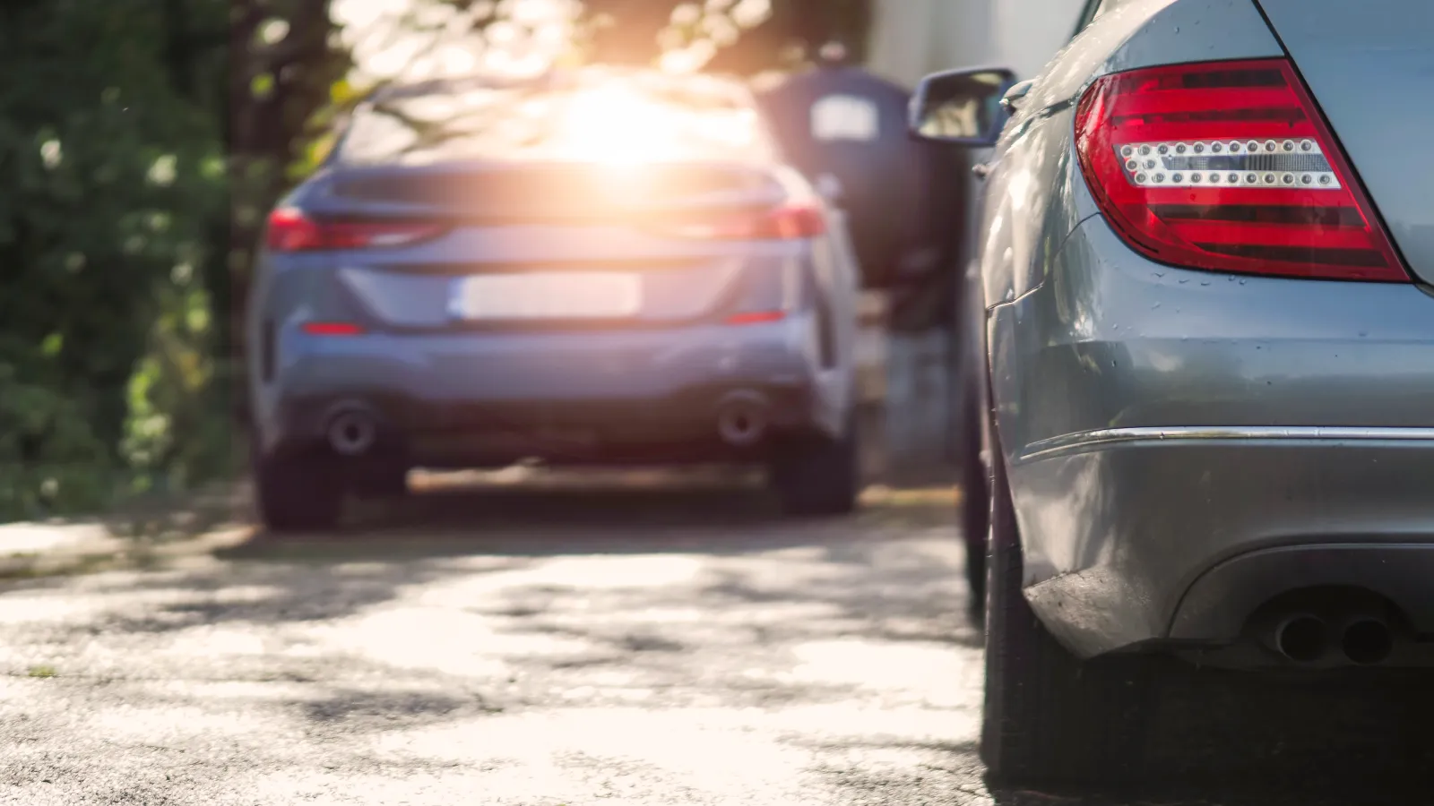 Silver and gray cars parked on a sunlit tree-lined street with bright glare in the background