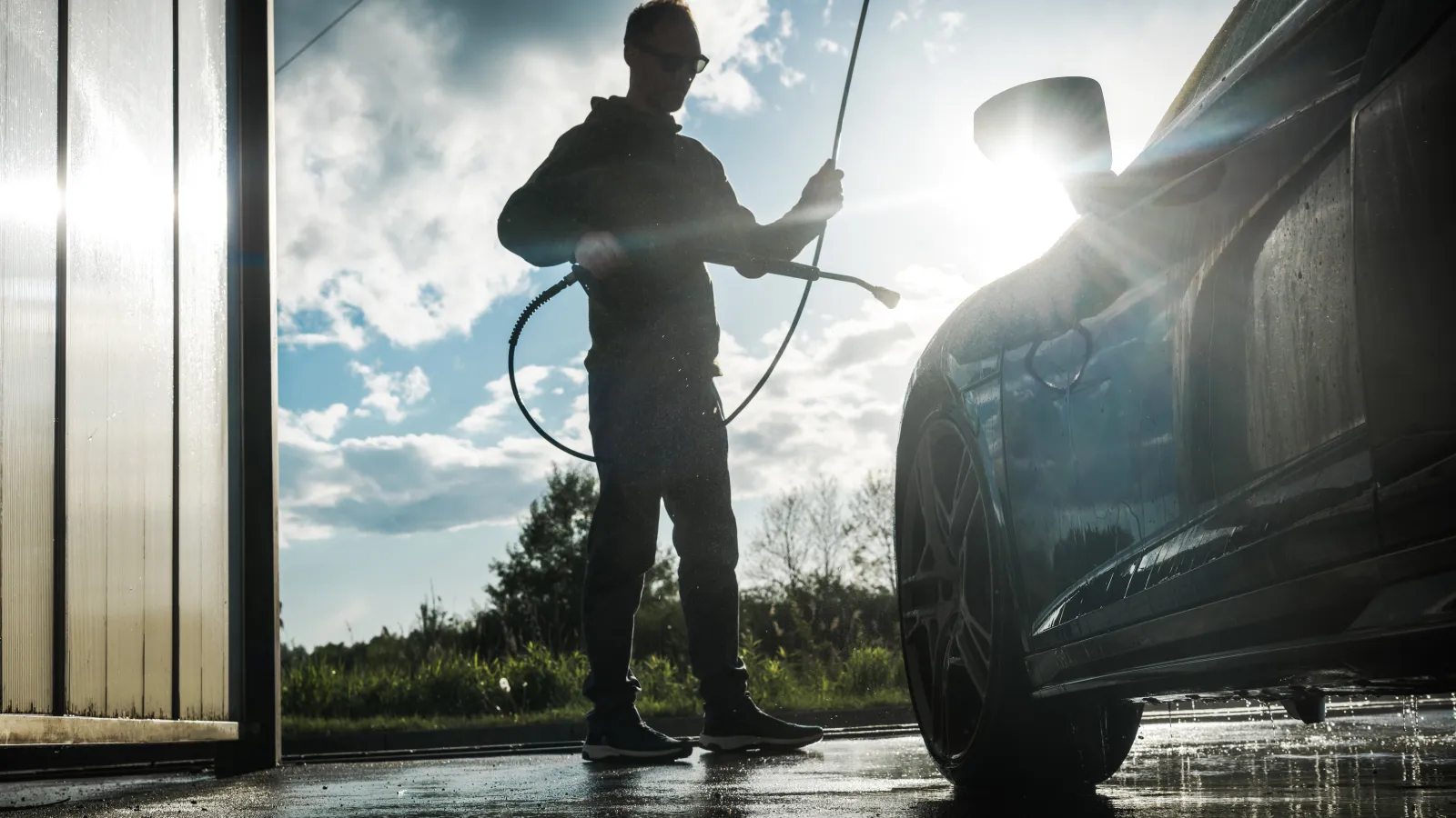 Person washing a car with a pressure washer outside on a sunny day with water on the ground.