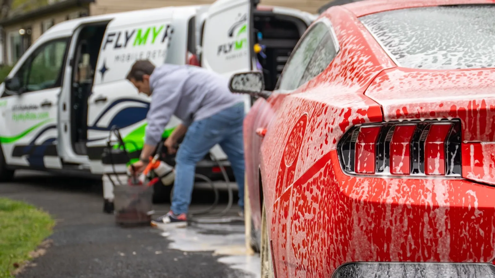 Red sports car covered in soap suds being washed by a person near a branded FlyMyCar mobile detailing van.