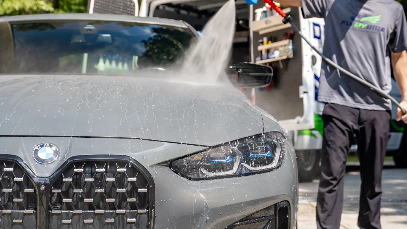 Man washing a gray BMW car outdoors with a high-pressure spray from a van in the background.