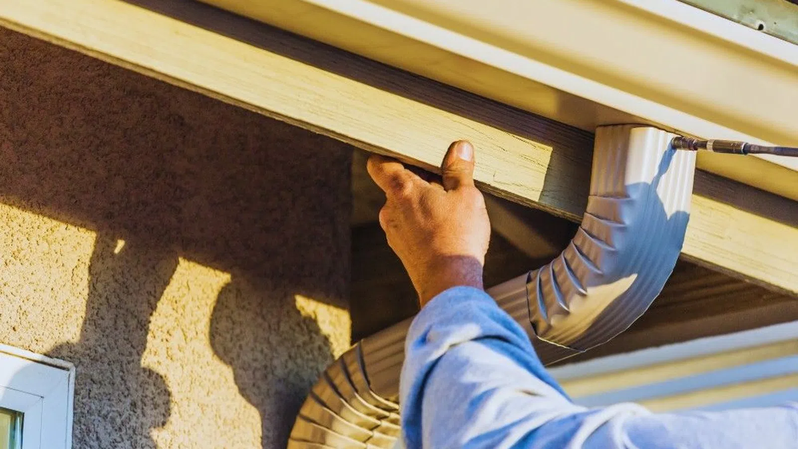 Worker installing wooden fascia board near white downspout gutter on house exterior under roof edge