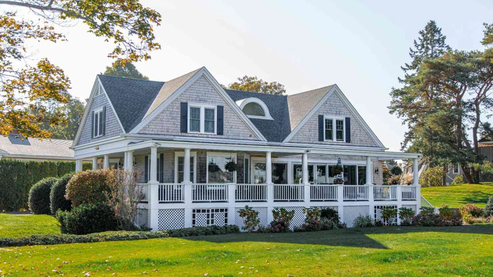 White coastal home with wraparound porch and manicured lawn under clear sky in suburban neighborhood