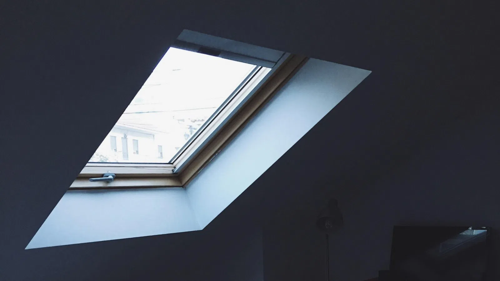 Dimly lit bedroom with a skylight, bed with open laptop, remote, and a desk with a heater and lamp.