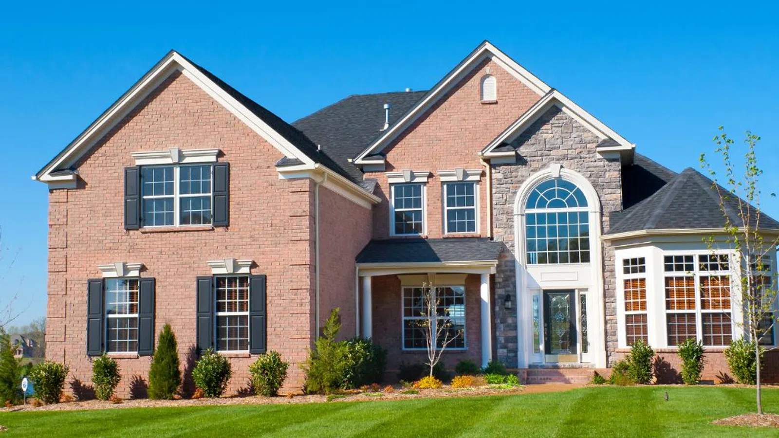 Large two-story brick and stone house with black shutters, tall arched window, and manicured green lawn on a sunny day