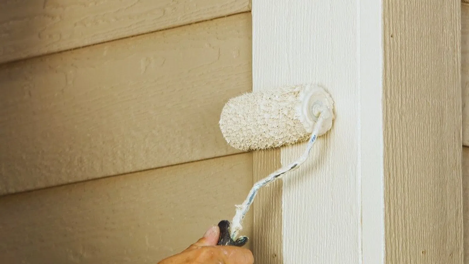 Hand painting exterior house wall trim white using paint roller on beige siding.