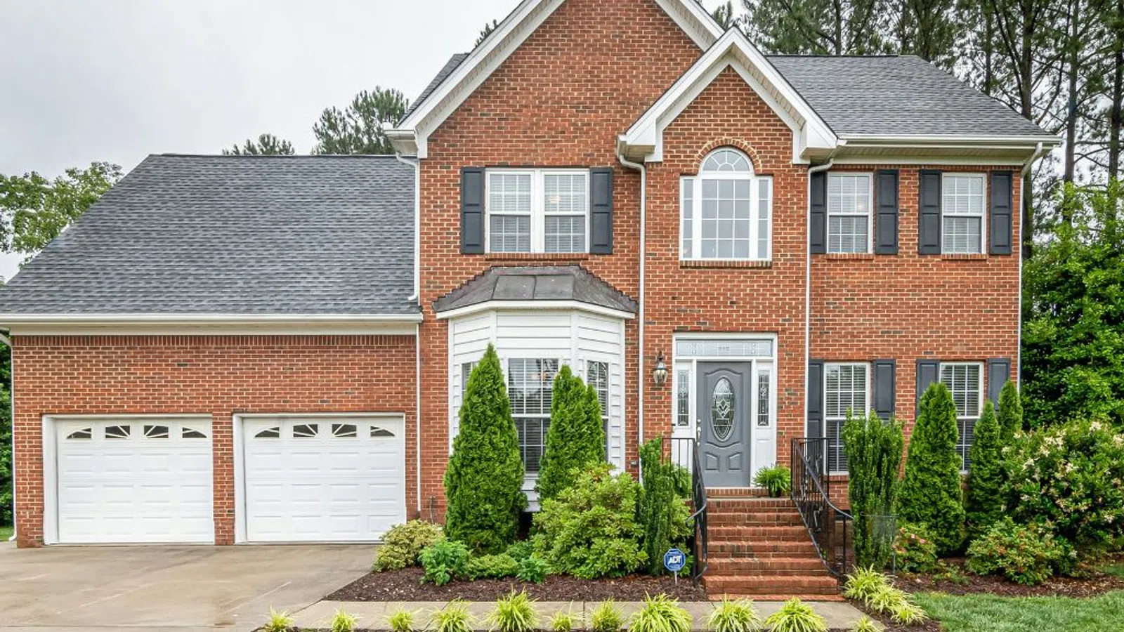 Two-story red brick house with white trim, gray roof, double garage, front steps, and well-maintained landscaping.