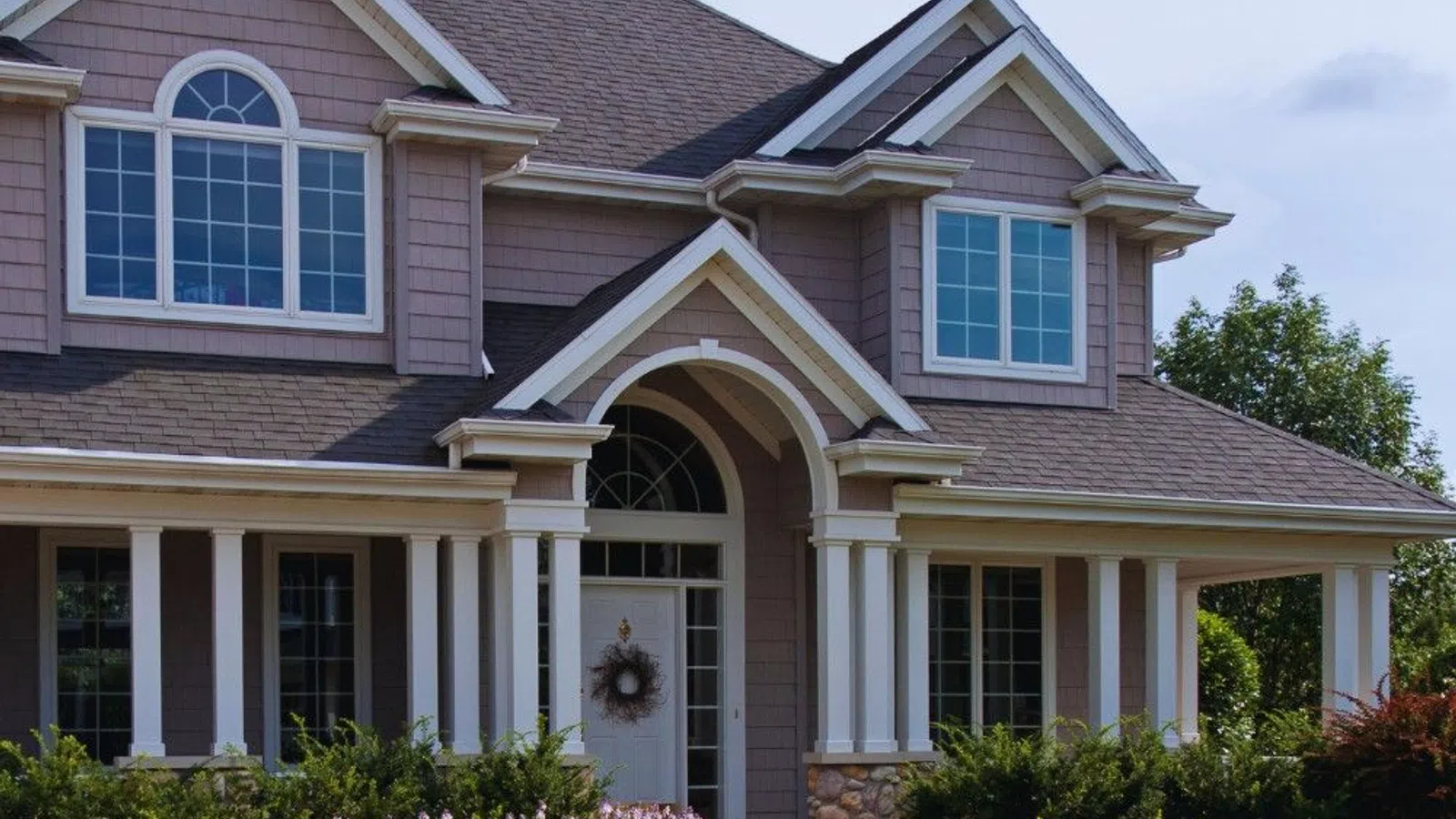 Two-story house with large windows, gable roof, white columns, stone accents, and landscaped garden under blue sky