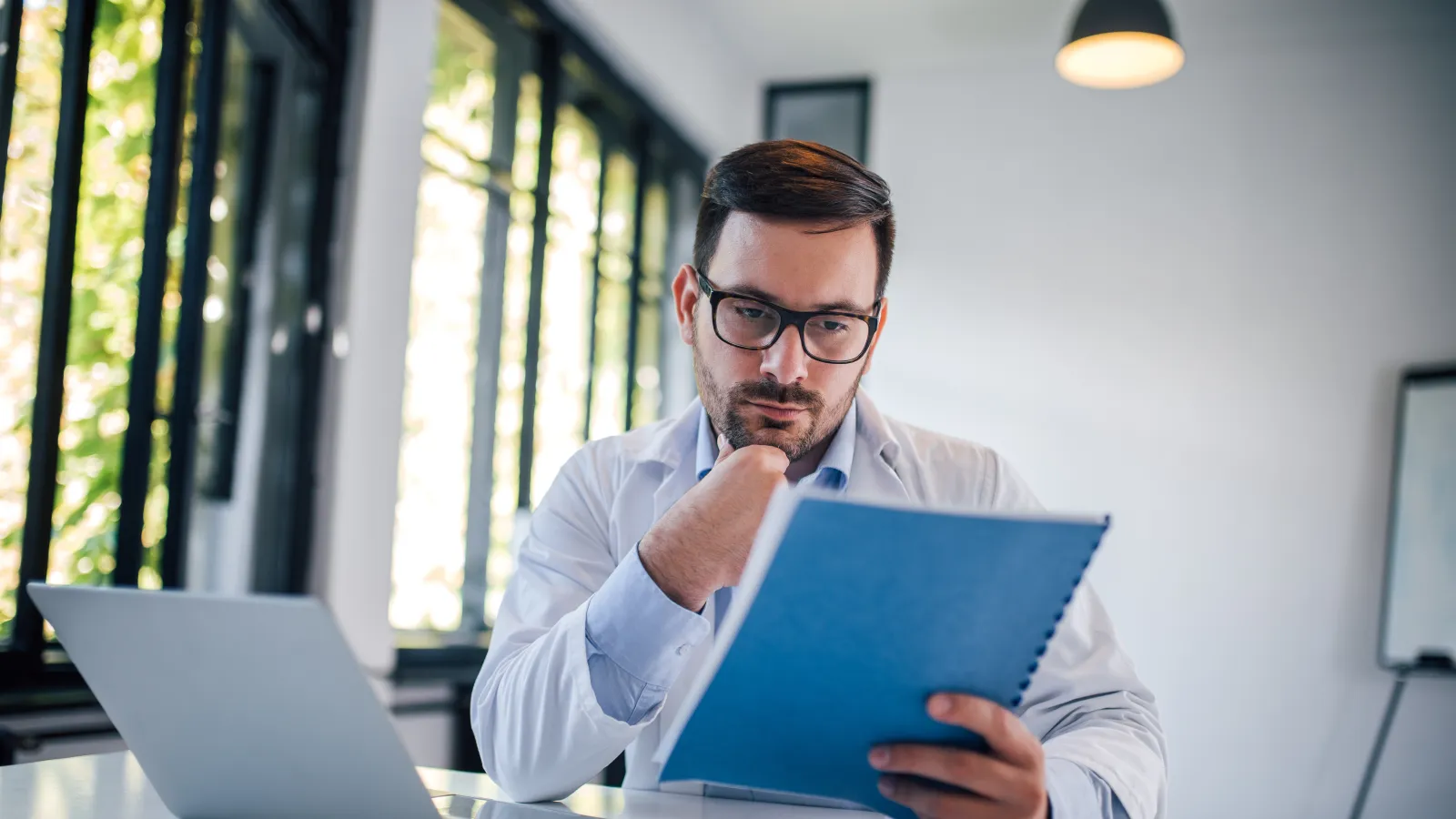 Male doctor in white coat reviewing patient file at desk with laptop and stethoscope in a bright office.