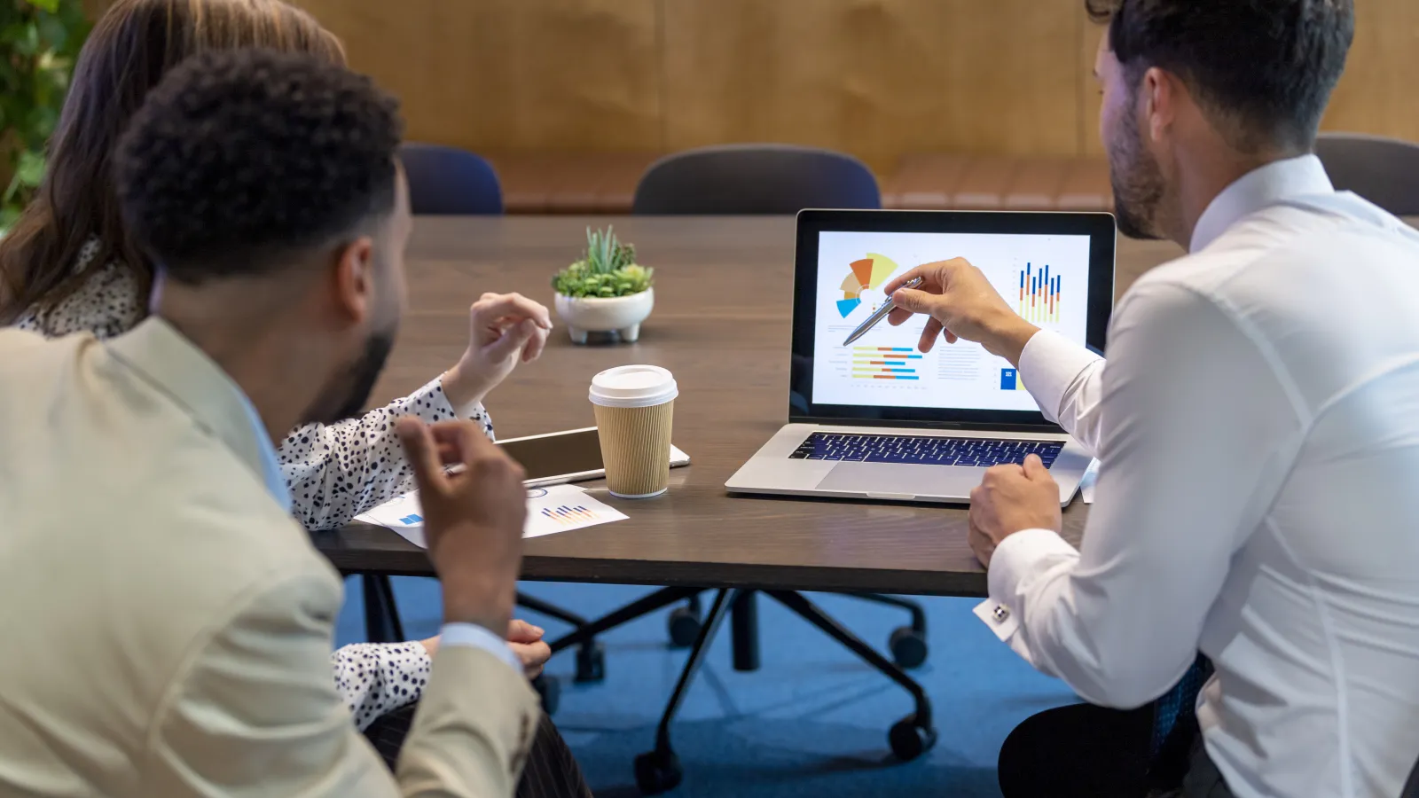 Three professionals discussing financial charts on a laptop during a meeting in a conference room.