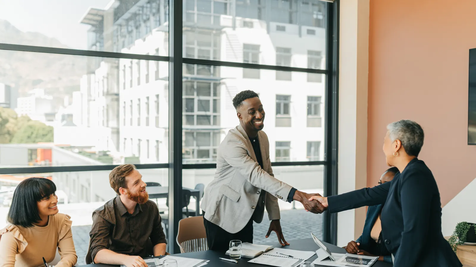 Diverse professionals shaking hands in a modern office meeting with colleagues taking notes and smiling.