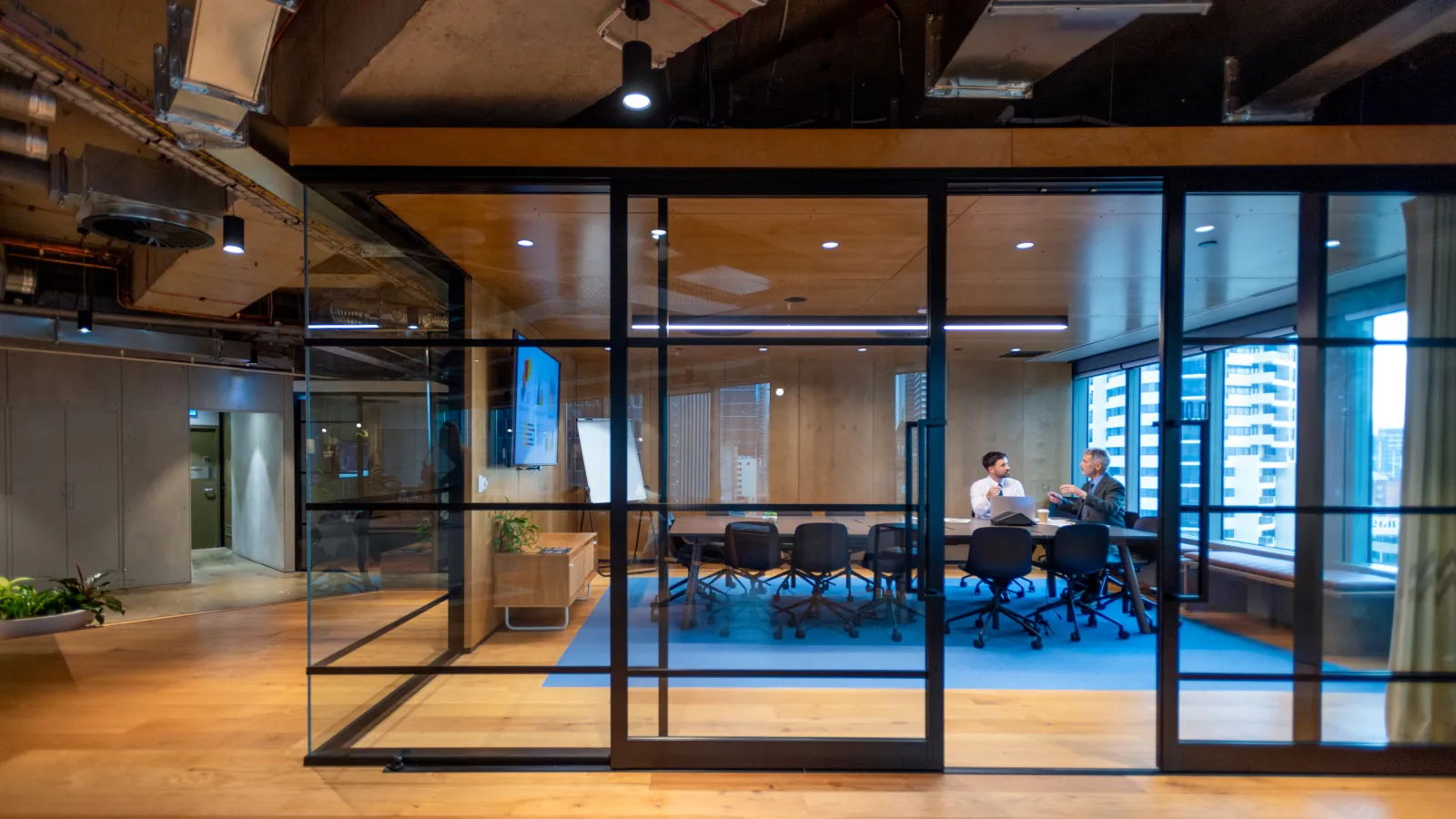 Modern office conference room with glass walls, wooden flooring, and two people discussing inside.