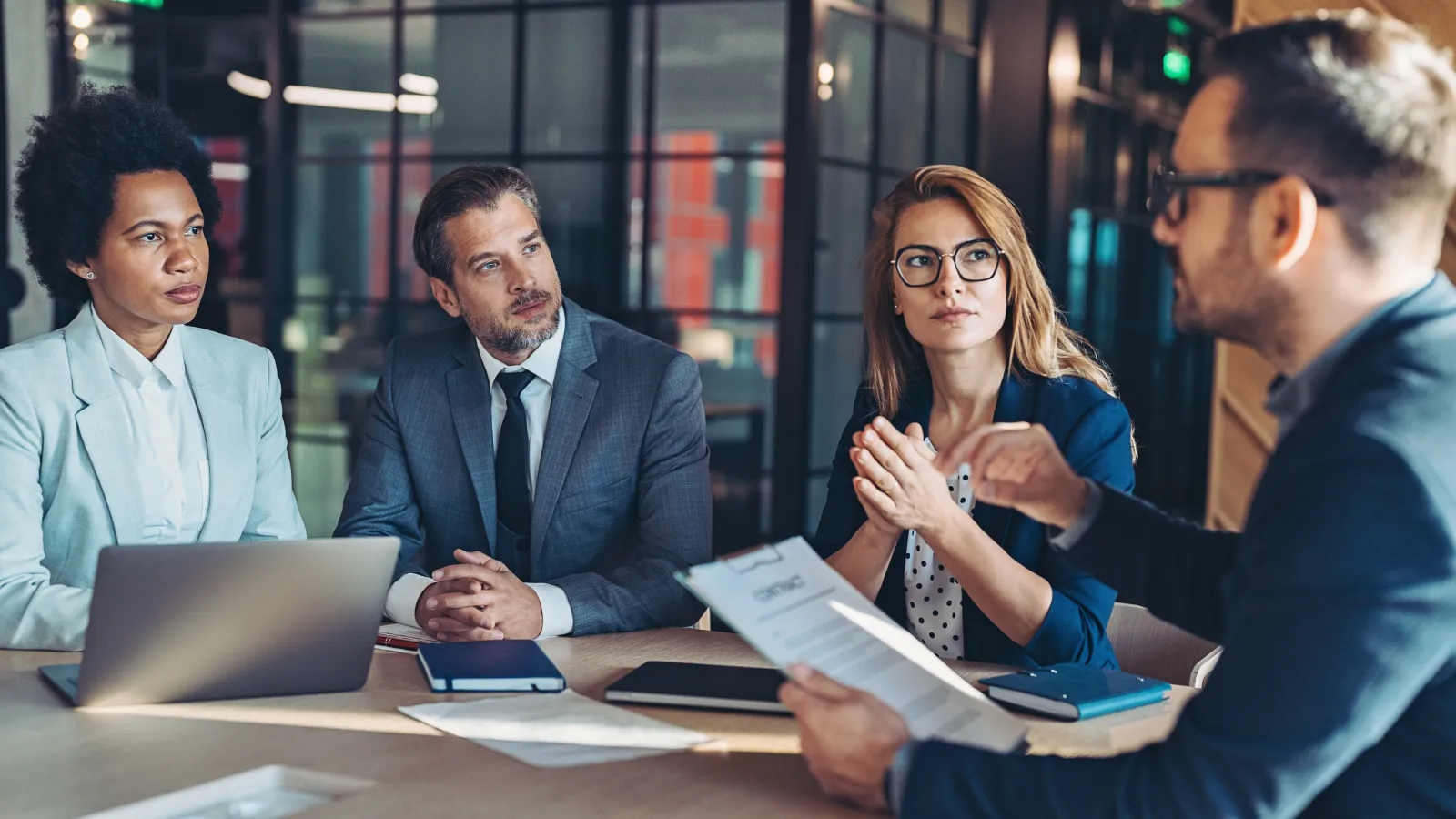 Four diverse professionals engaged in a serious business meeting around a wooden table with documents and laptops.