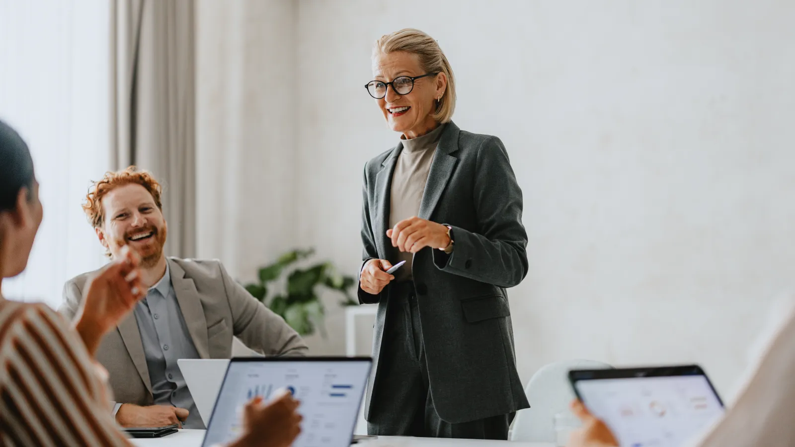 Smiling businesswoman in gray suit leading an engaging meeting with colleagues using laptops in a bright office.