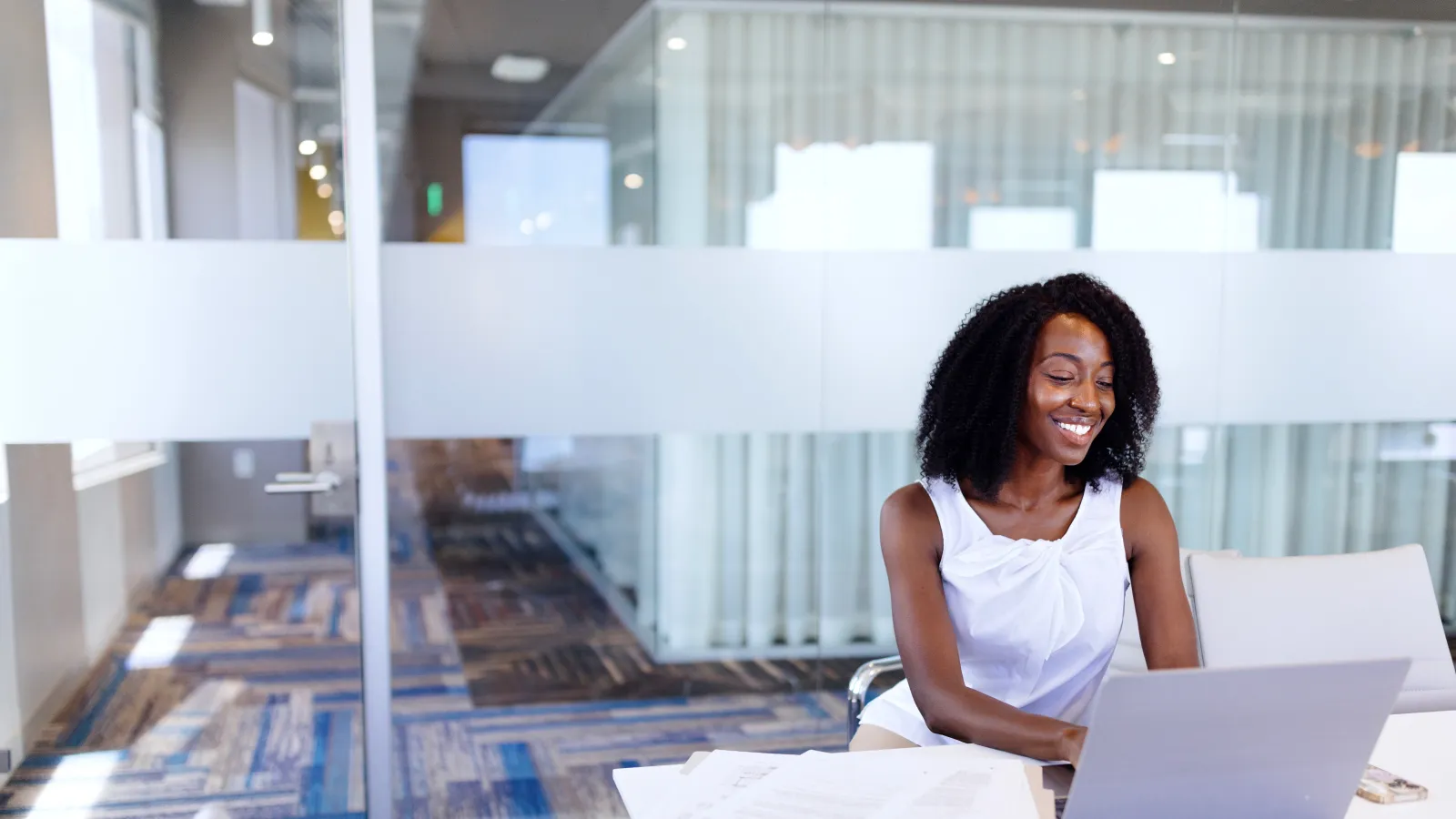 Smiling woman working on laptop at desk with documents in modern office with glass walls and blue carpet.