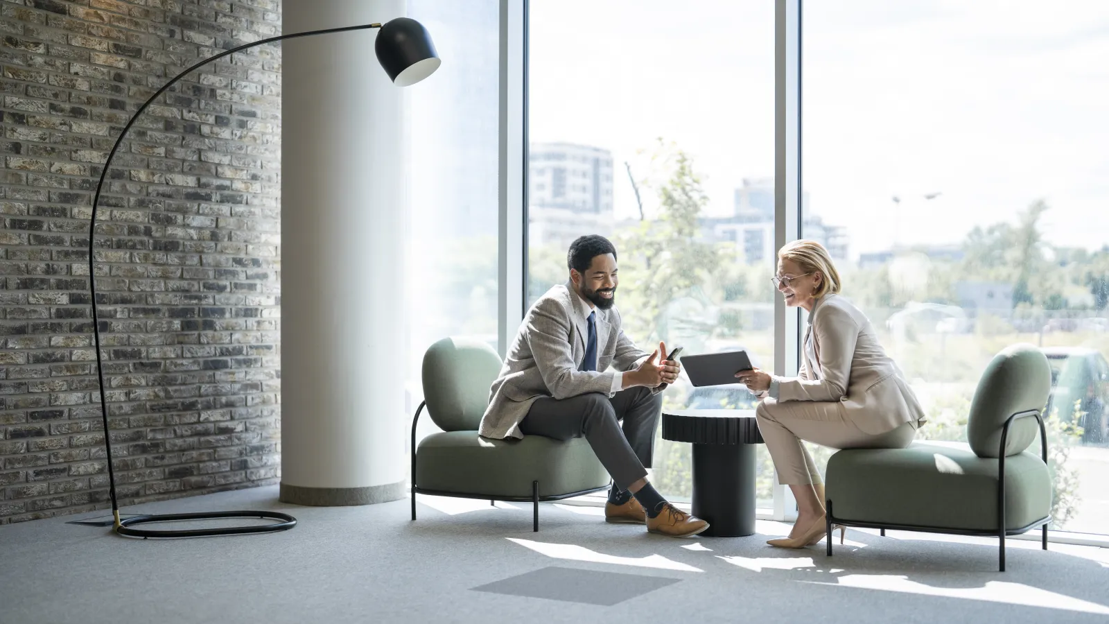 Two business professionals discussing work on a tablet in a modern office with large windows and green chairs.