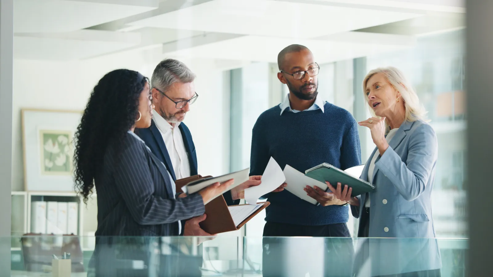 Four diverse professionals discussing documents in a modern office with glass walls and natural light