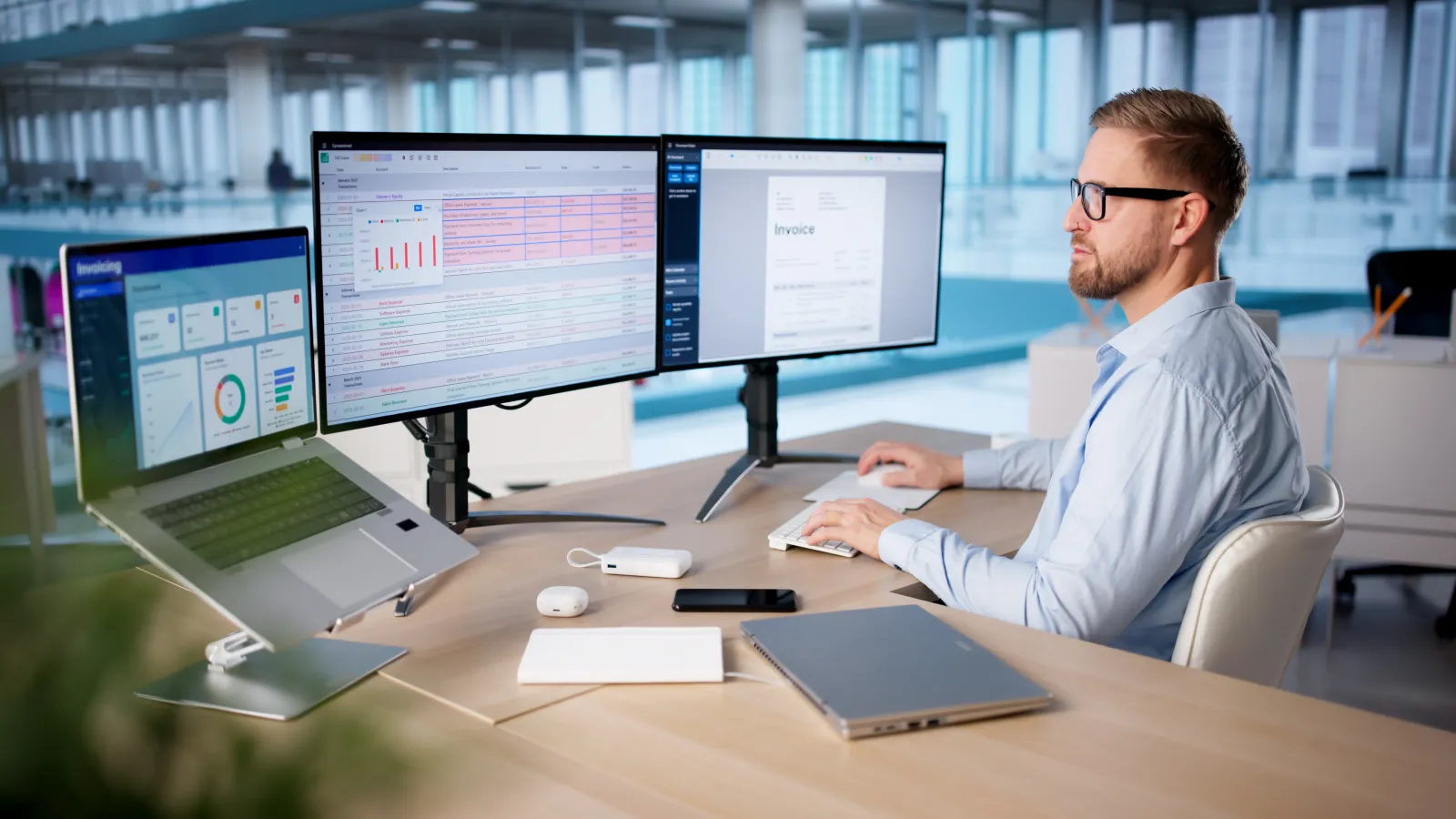 Man in office using a computer with dual monitors displaying data and invoices in a modern workspace.
