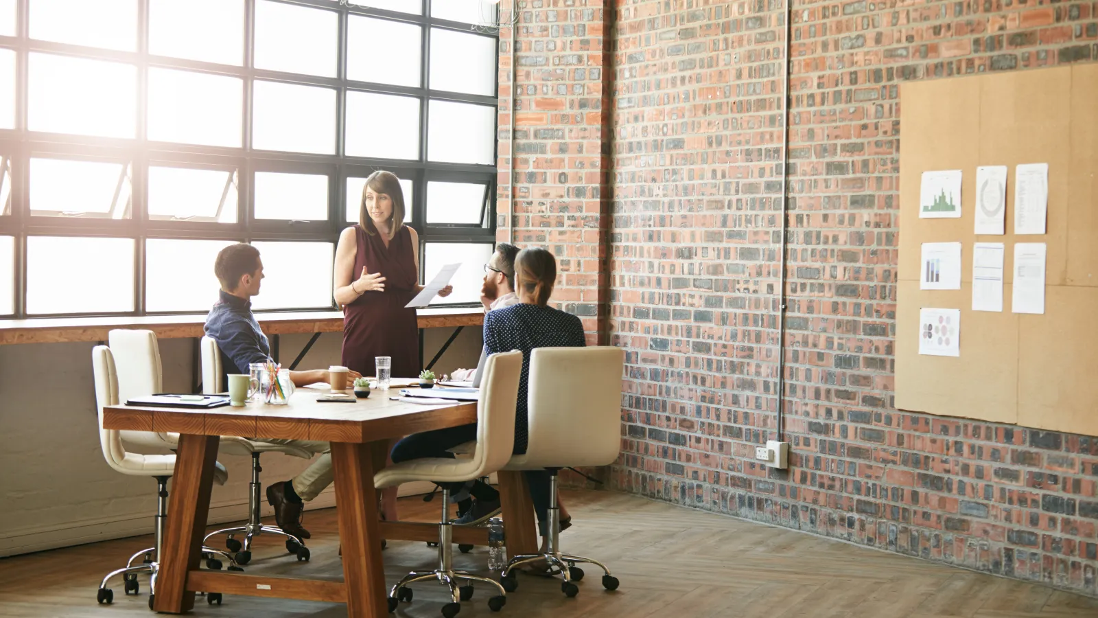 Team meeting in modern office with brick walls and large windows, woman presenting papers to colleagues.