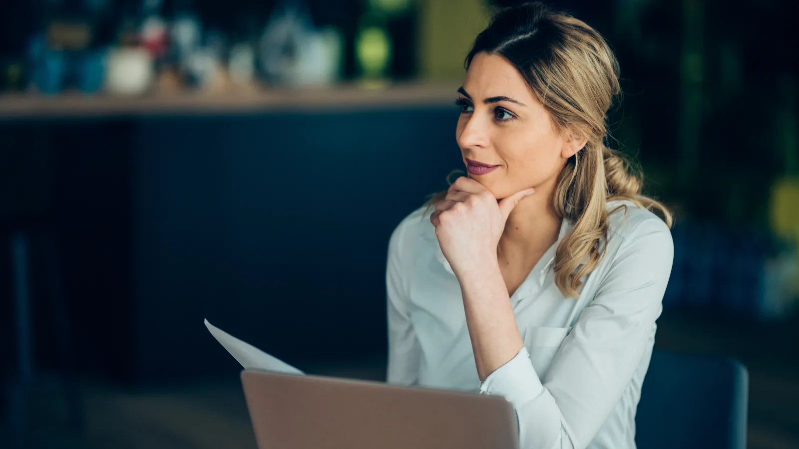 Woman in white shirt working on laptop and holding paper, looking thoughtfully to side indoors