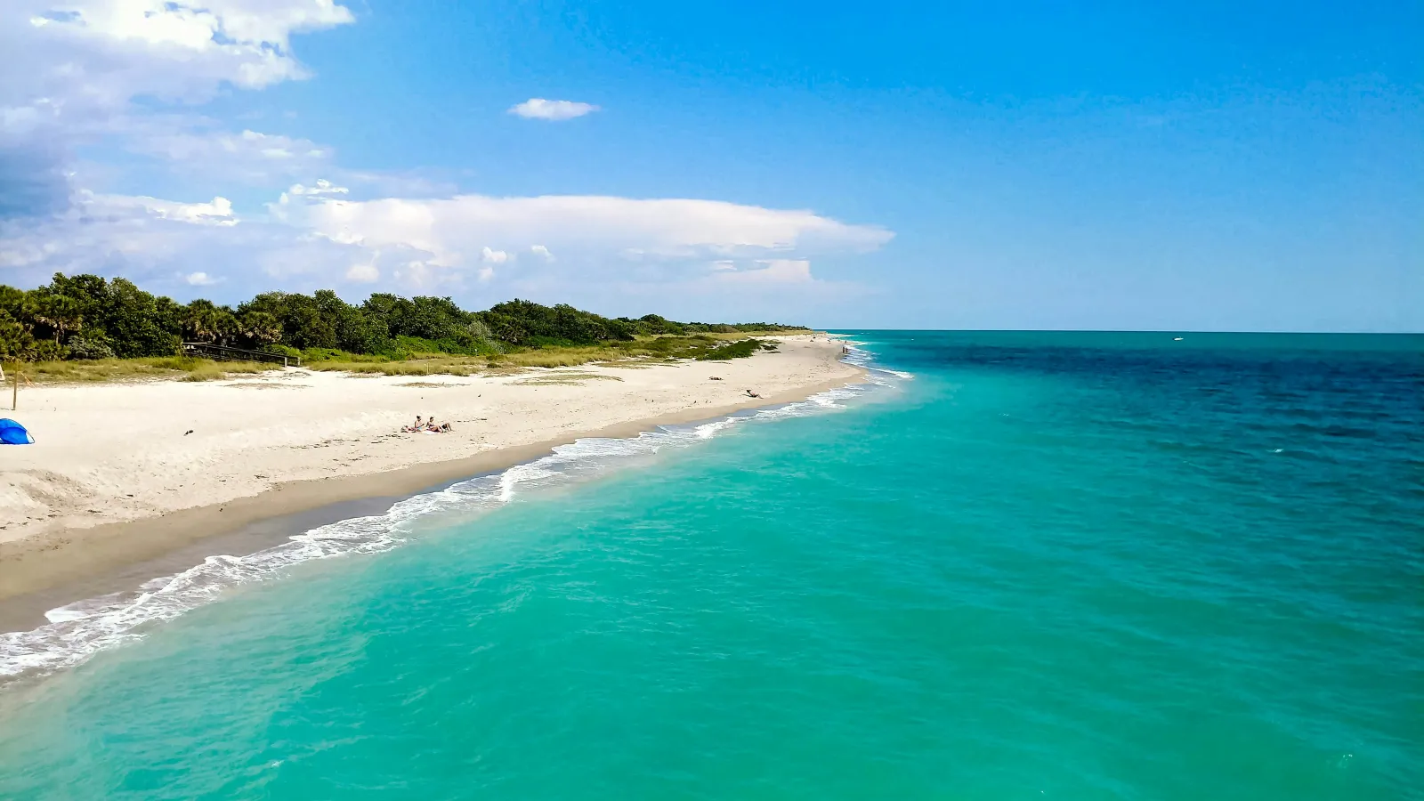 Pristine sandy beach with clear turquoise water, green vegetation, and a bright blue sky with scattered clouds.