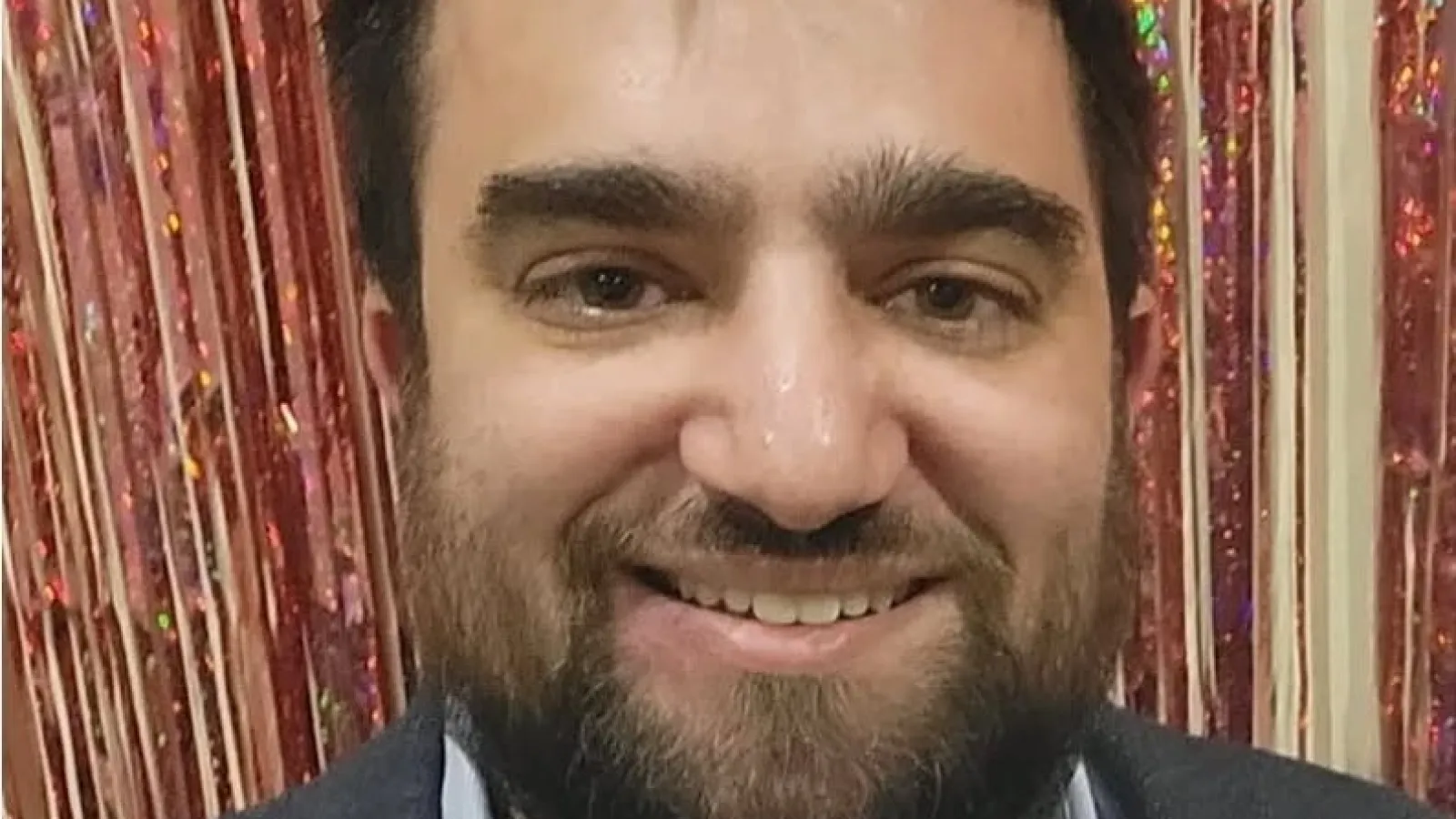 Smiling man with beard wearing suit jacket and light blue shirt in front of a shiny pink curtain background