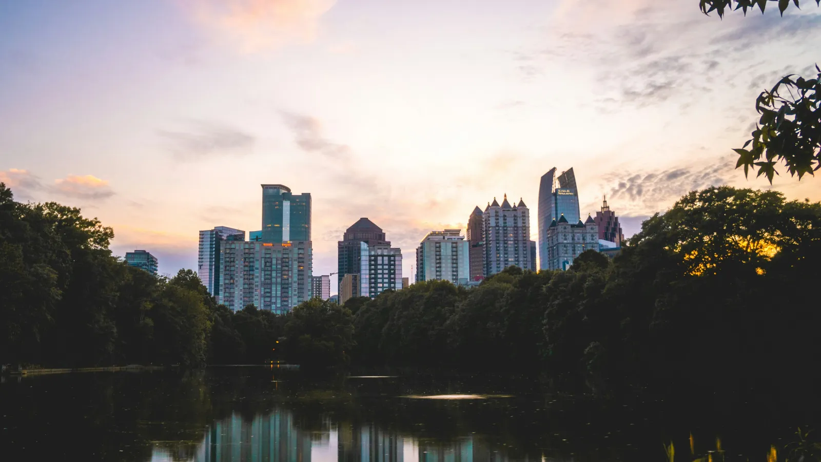 City skyline at sunset with tall buildings reflected in the calm water surrounded by trees.