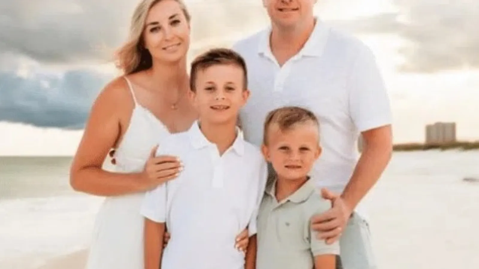 Family of four posing on a sandy beach with cloudy sky in the background, wearing casual summer clothes.