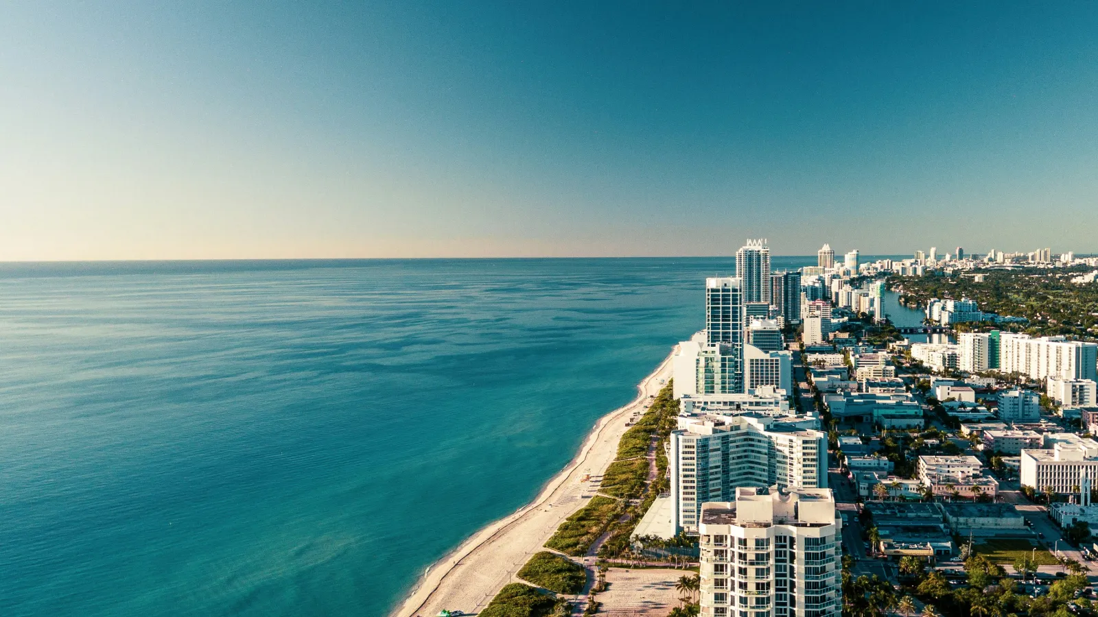 Aerial view of a coastal city with tall buildings lining a sandy beach and calm blue ocean under a clear sky.