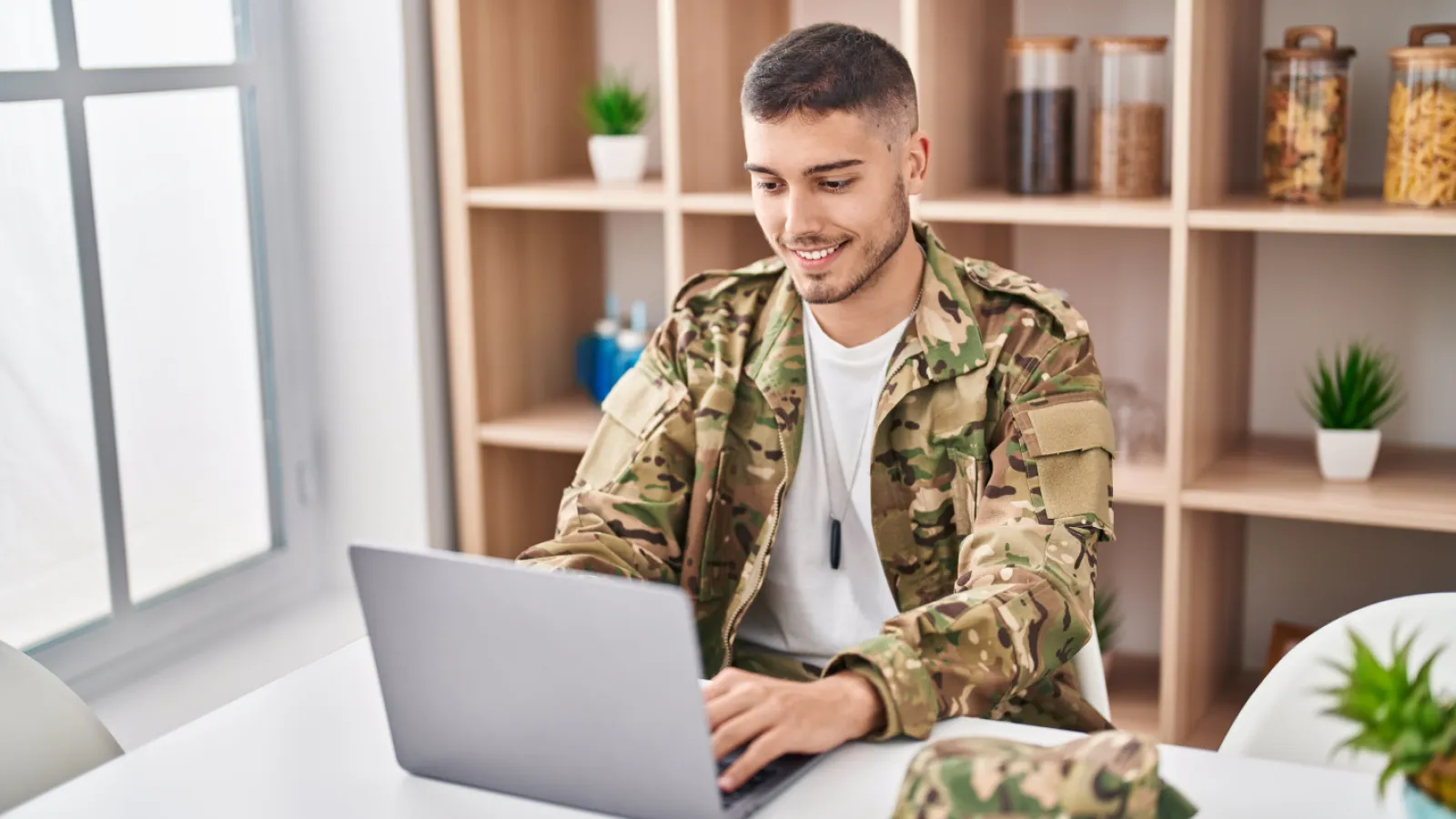 Young military man in camouflage uniform using laptop and smiling while sitting at white desk indoors.