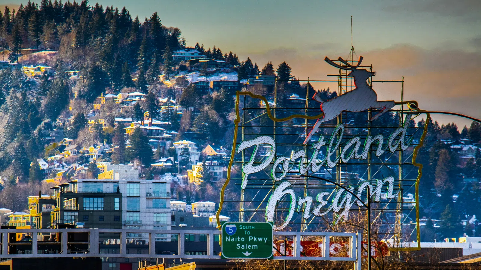 Iconic Portland Oregon sign with a white stag against a backdrop of forested hills and city buildings at sunset