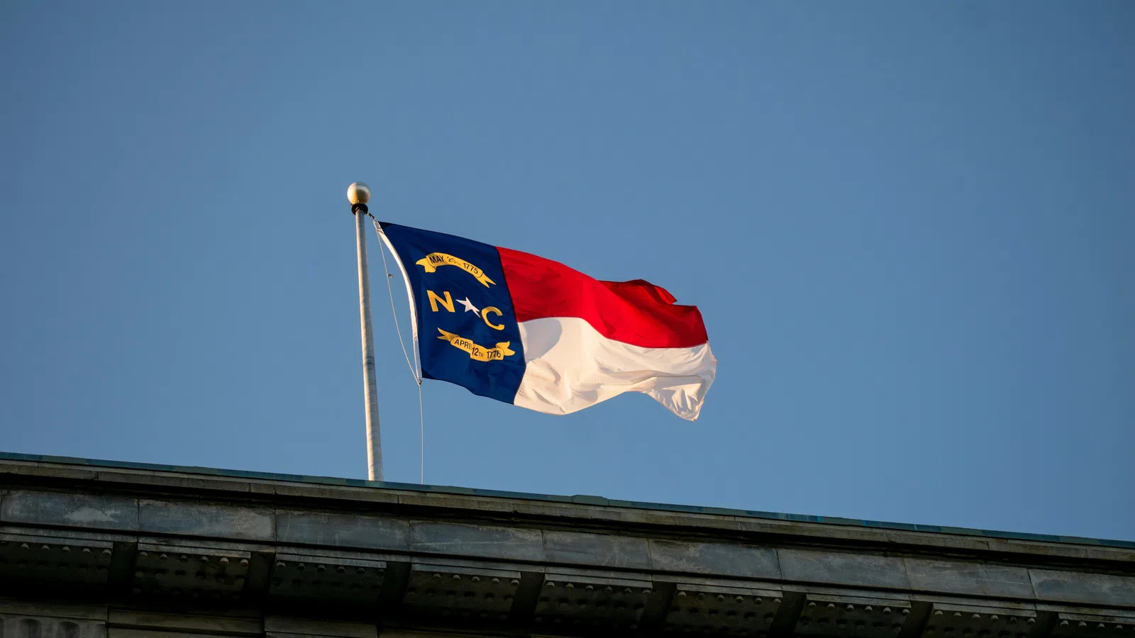 North Carolina state flag waving on a flagpole above a building against a clear blue sky.