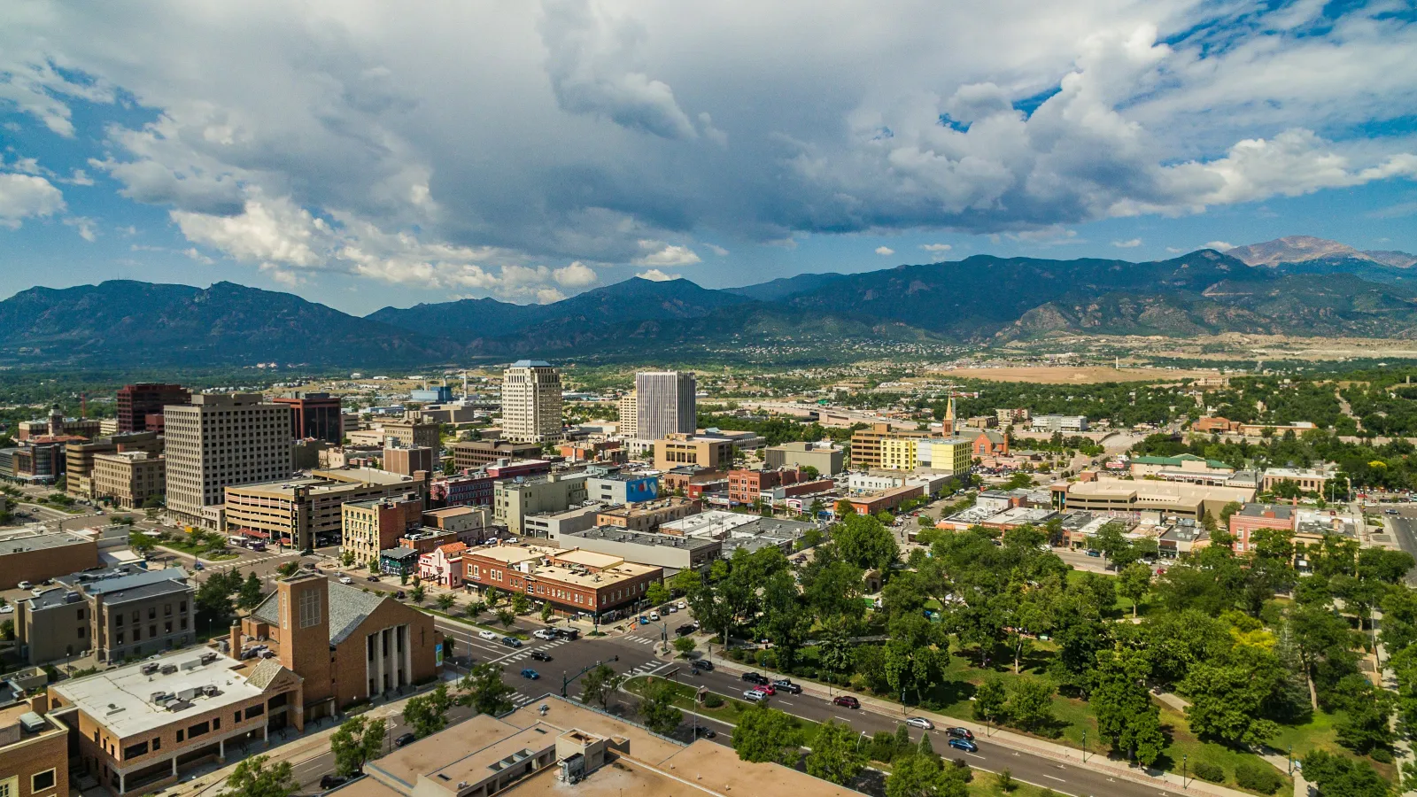 Aerial view of a city with mid-rise buildings, green parks, and mountains in the background under a partly cloudy sky.