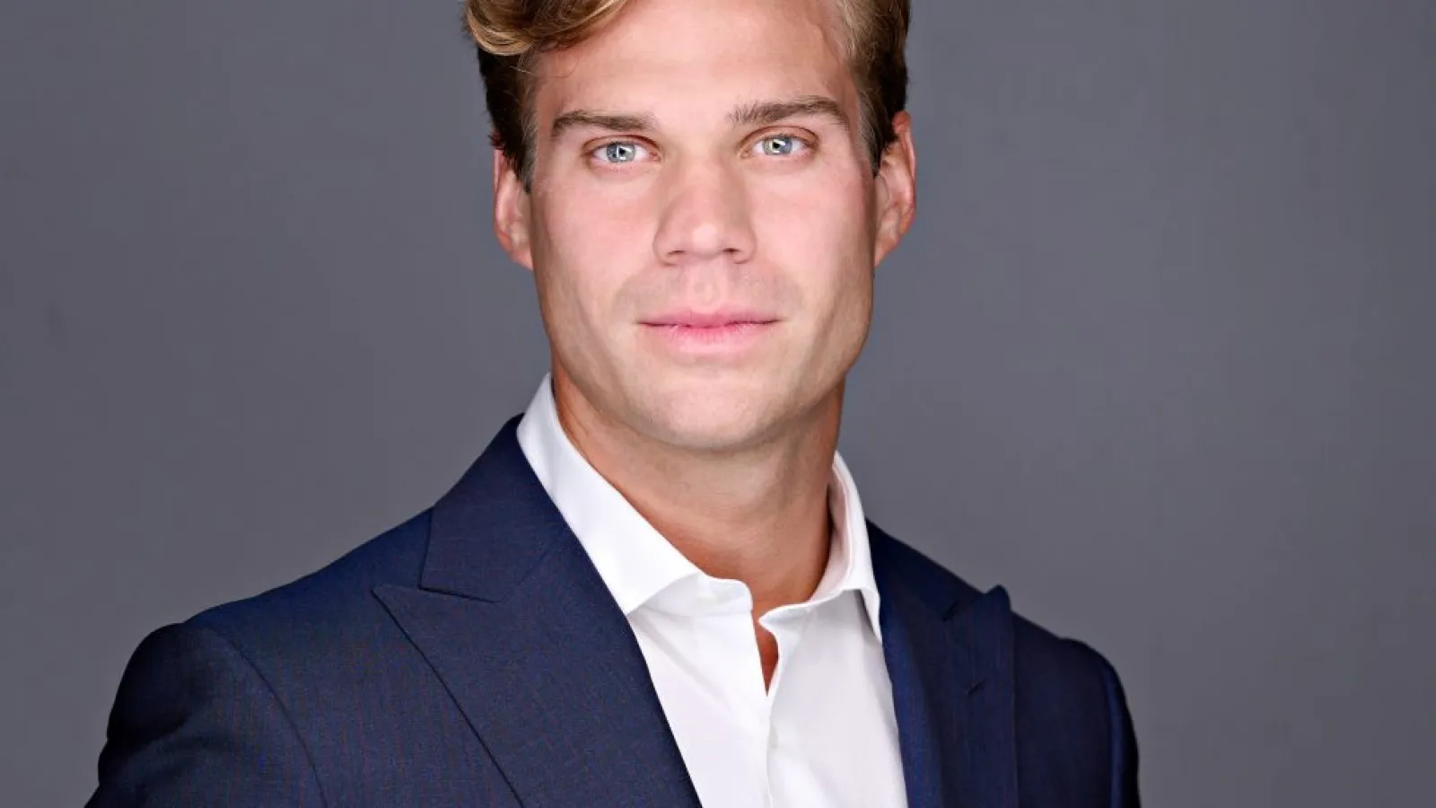 Professional man with blond hair wearing a navy blue suit and white shirt against a gray background.