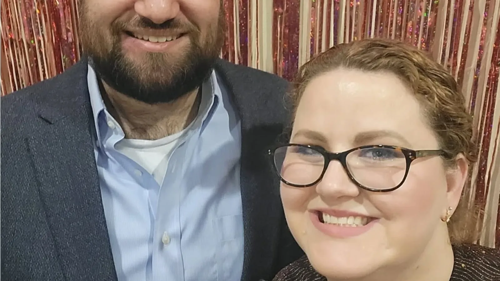 Smiling man in blue shirt and blazer and woman with glasses posing in front of shiny pink curtain backdrop.