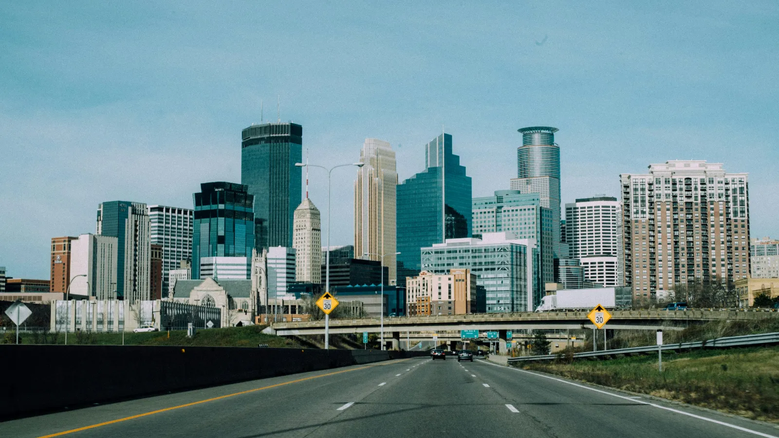 Wide highway leading into a modern city skyline with tall skyscrapers under a clear blue sky.