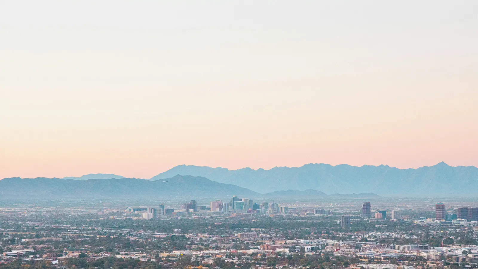 Panoramic cityscape with urban buildings and distant mountains under a soft pastel sky at sunset.