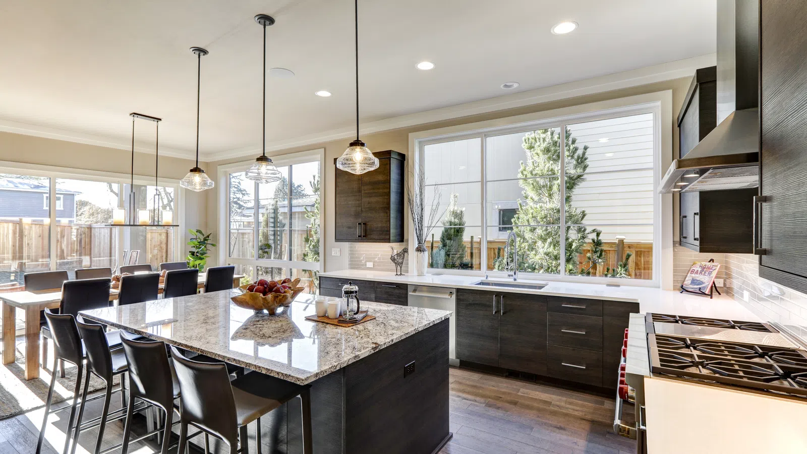 Modern kitchen with large granite island, dark cabinets, pendant lights, dining area, and large windows letting in natural light