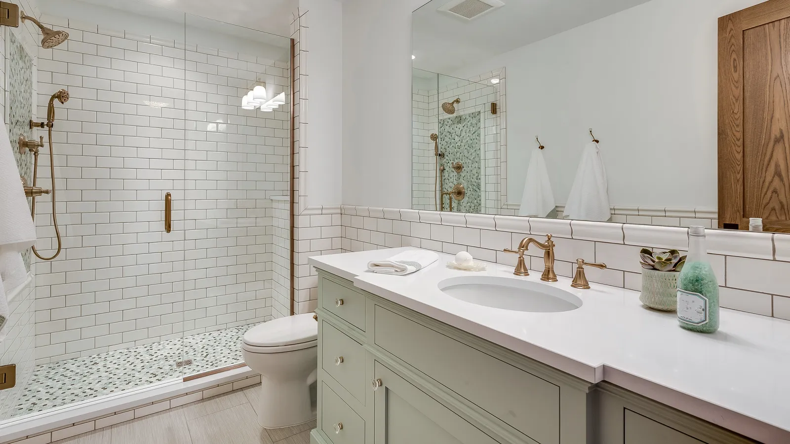 Modern bathroom with glass shower, white subway tiles, green vanity, and brass fixtures with mirror above.