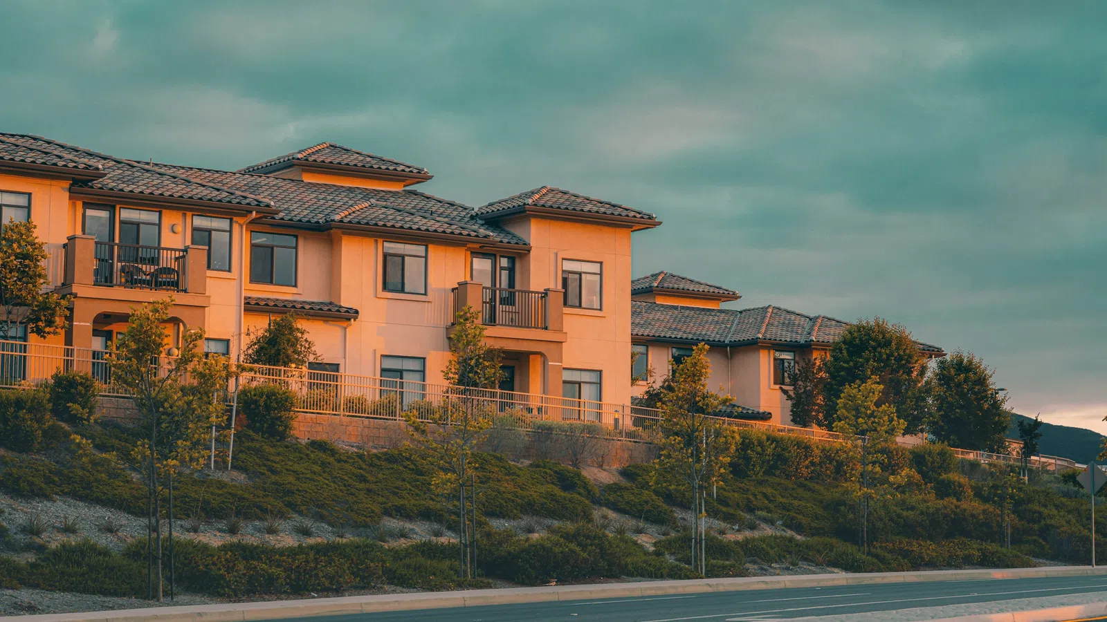 Modern multi-unit residential buildings with tiled roofs along a landscaped street at sunset under cloudy sky