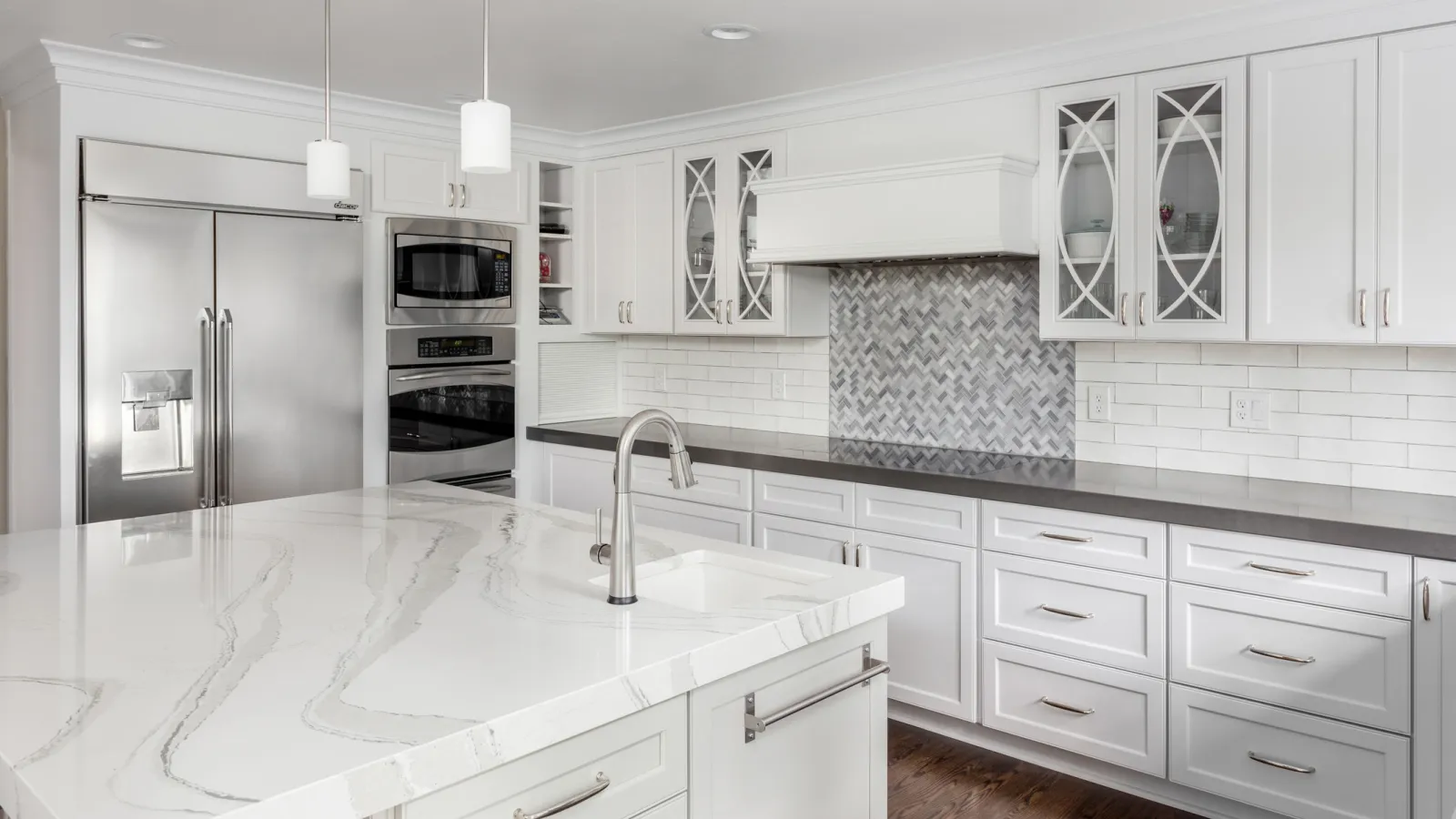 Modern white kitchen with marble island, stainless steel appliances, and gray backsplash with pendant lights.