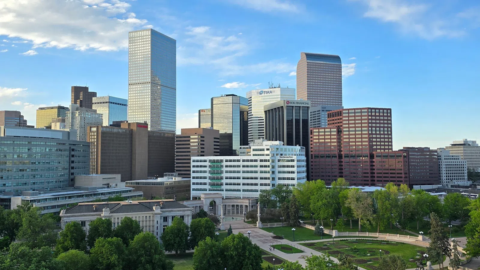 Downtown city skyline with modern skyscrapers and a green park under a partly cloudy blue sky