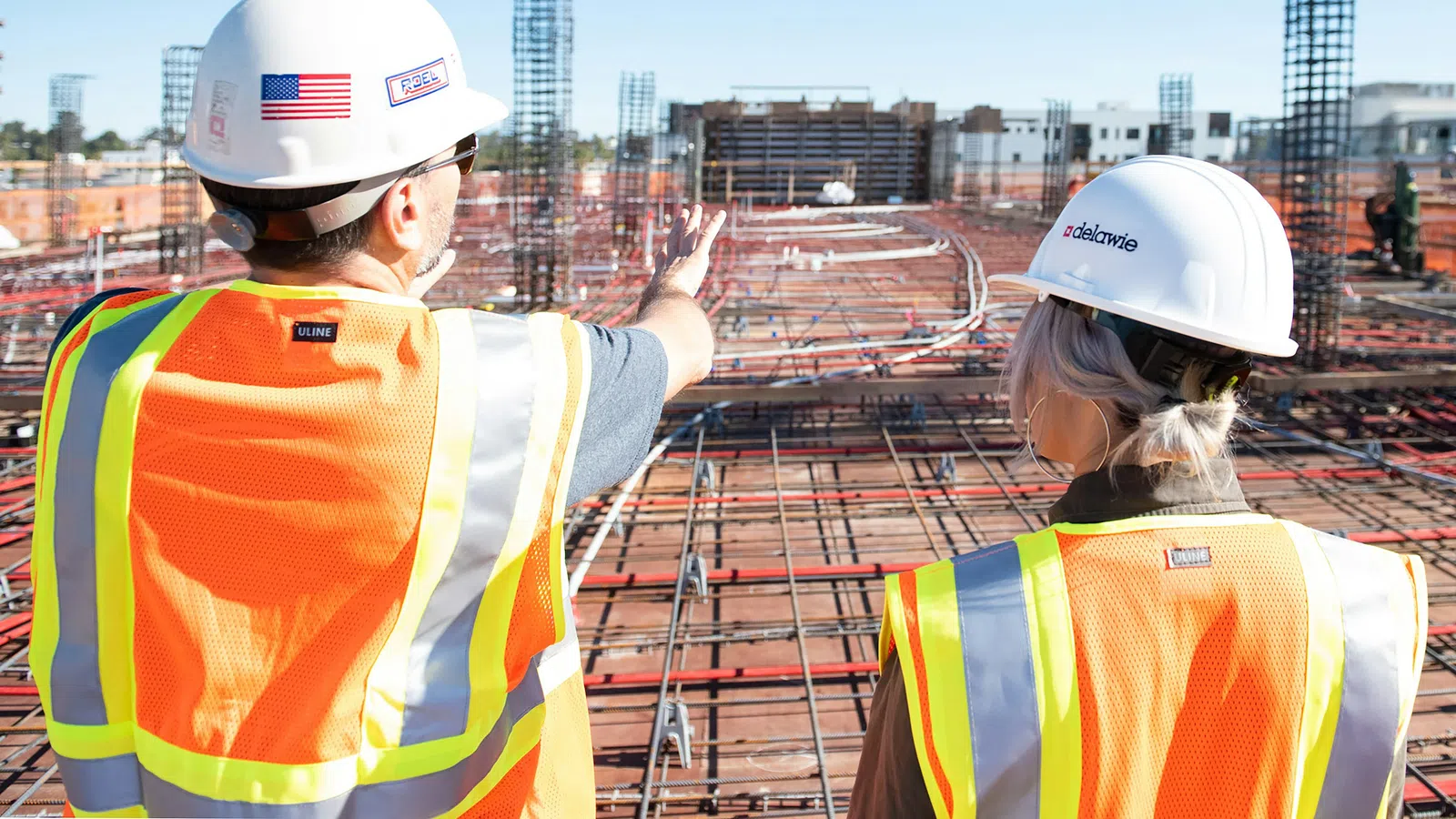 Two construction workers in safety gear discuss progress on a steel-reinforced building foundation under a clear sky.