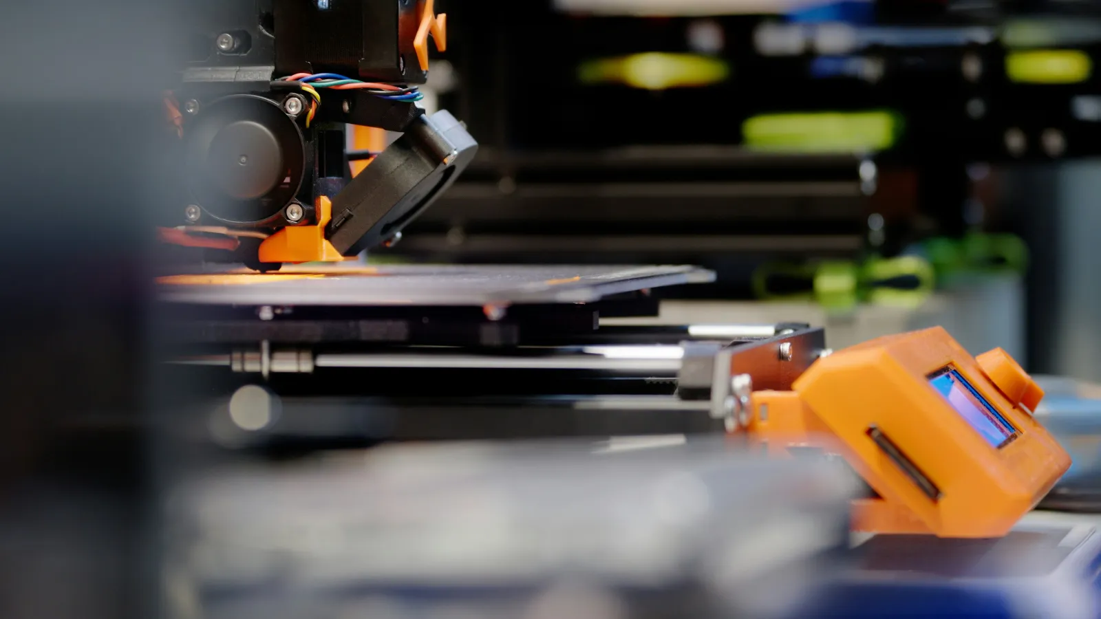 Close-up of a 3D printer in operation with orange components and a digital screen in a workshop setting
