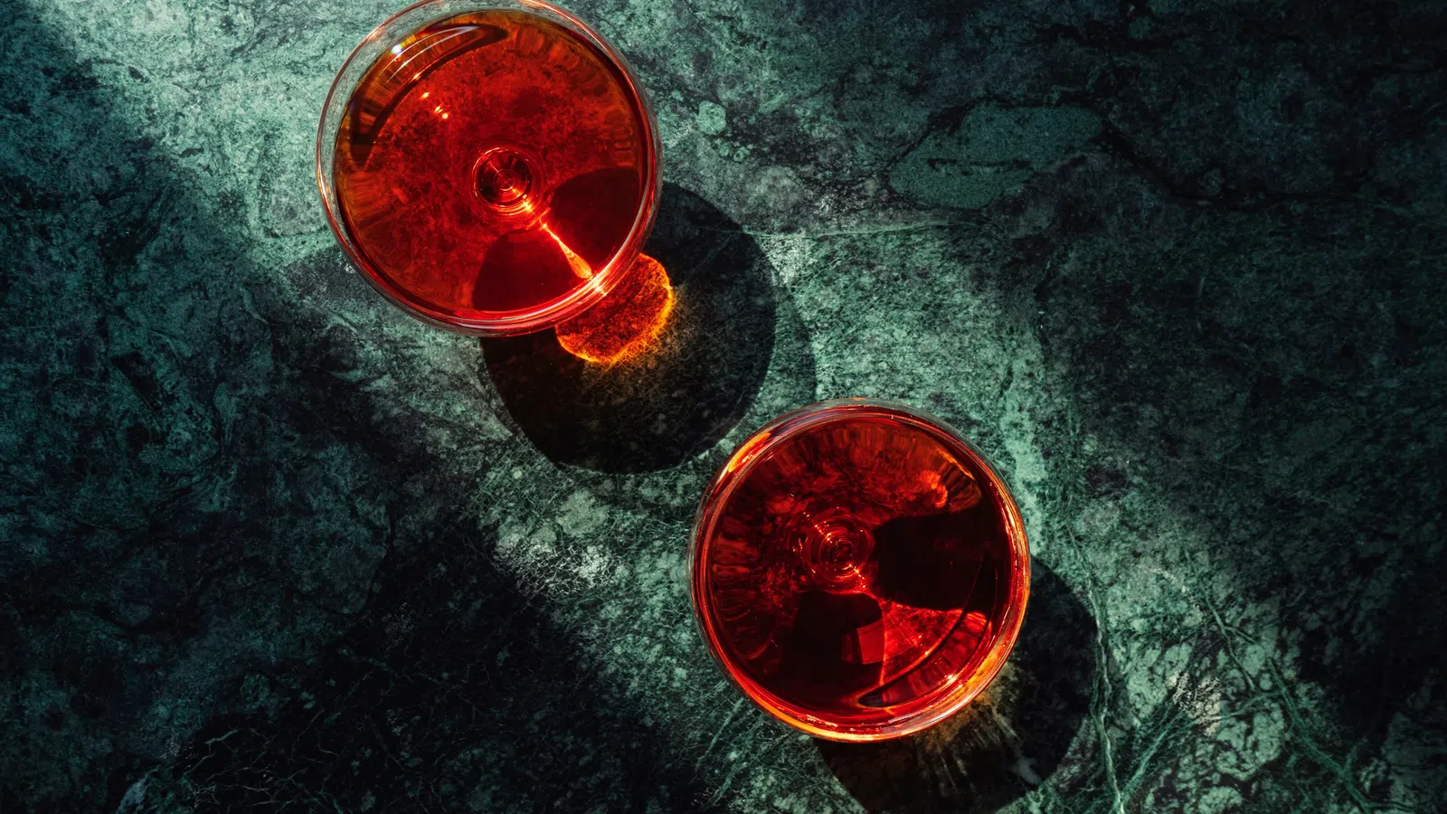 Two glasses of red wine casting shadows on a textured dark green marble surface in natural light.