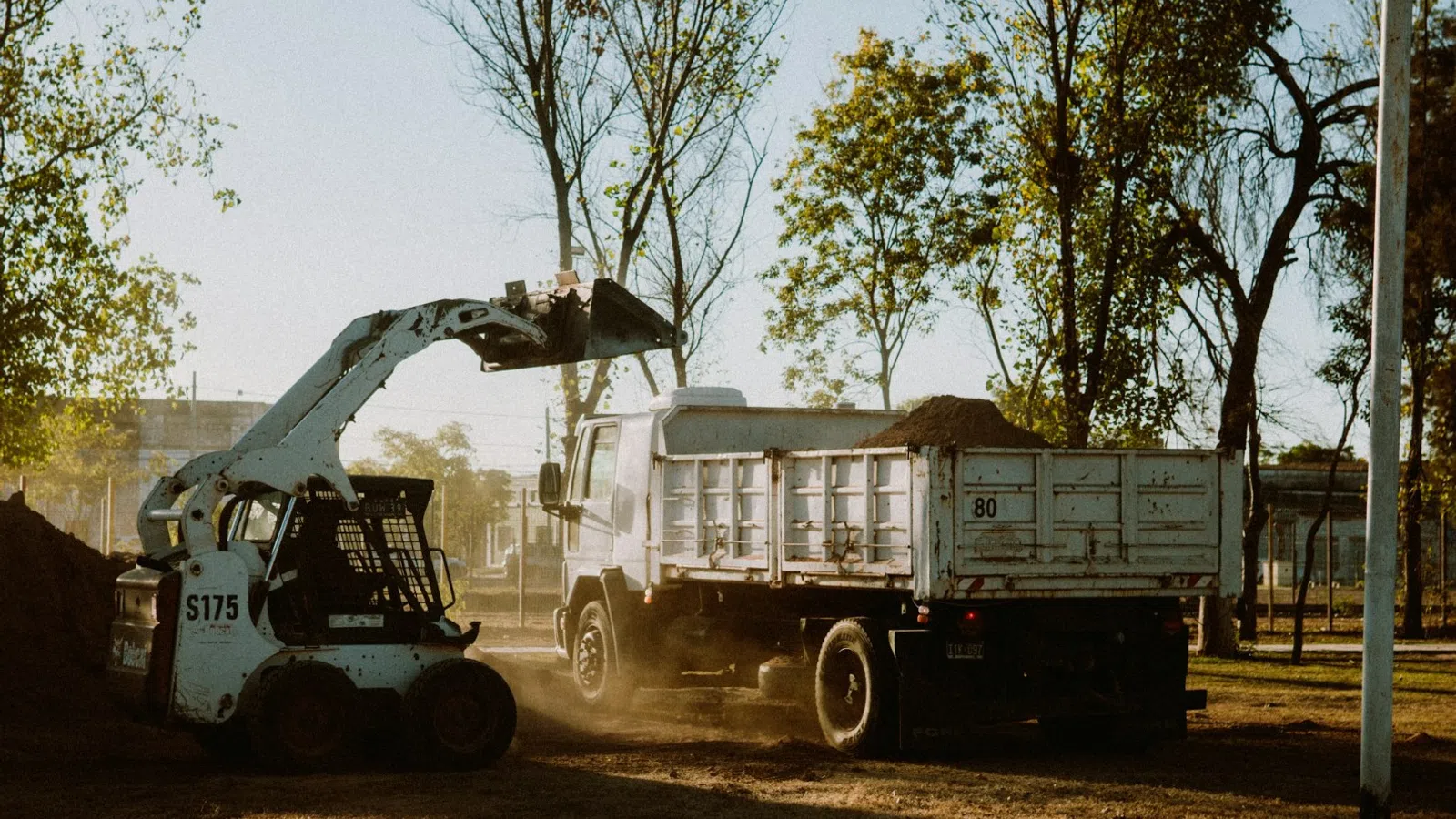 Loader filling a dump truck with dirt at a construction site surrounded by trees during daylight.