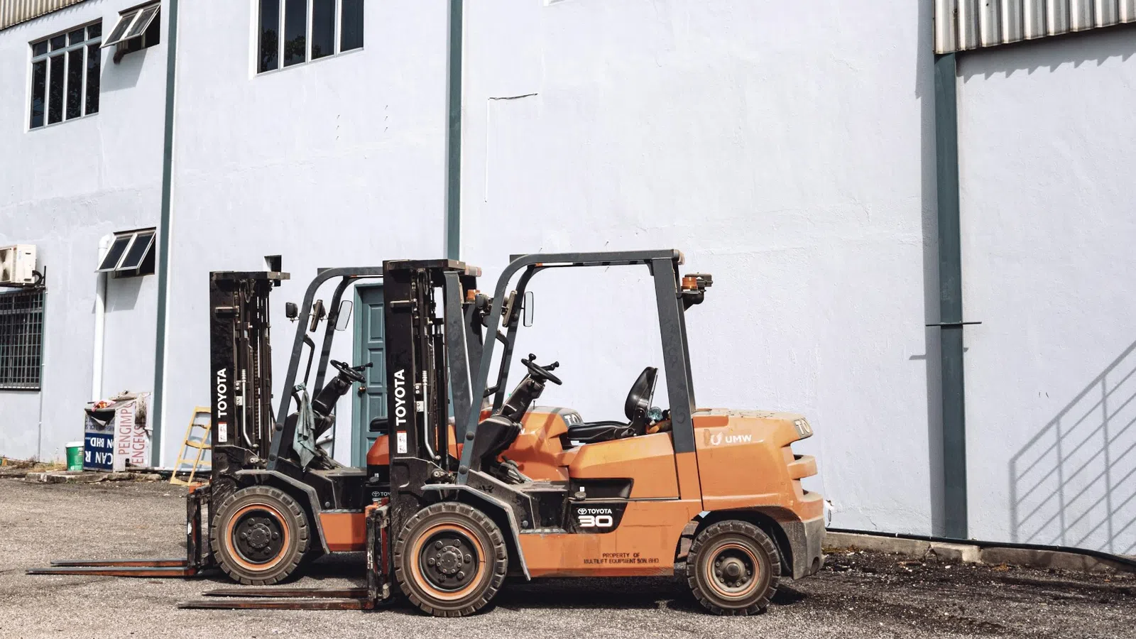 Two orange Toyota forklifts parked side by side outside a white industrial building on a sunny day
