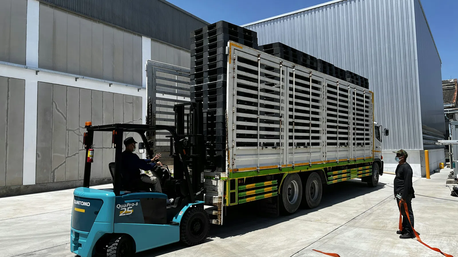 Forklift loading black crates onto a large white transport truck next to a warehouse under clear blue sky.