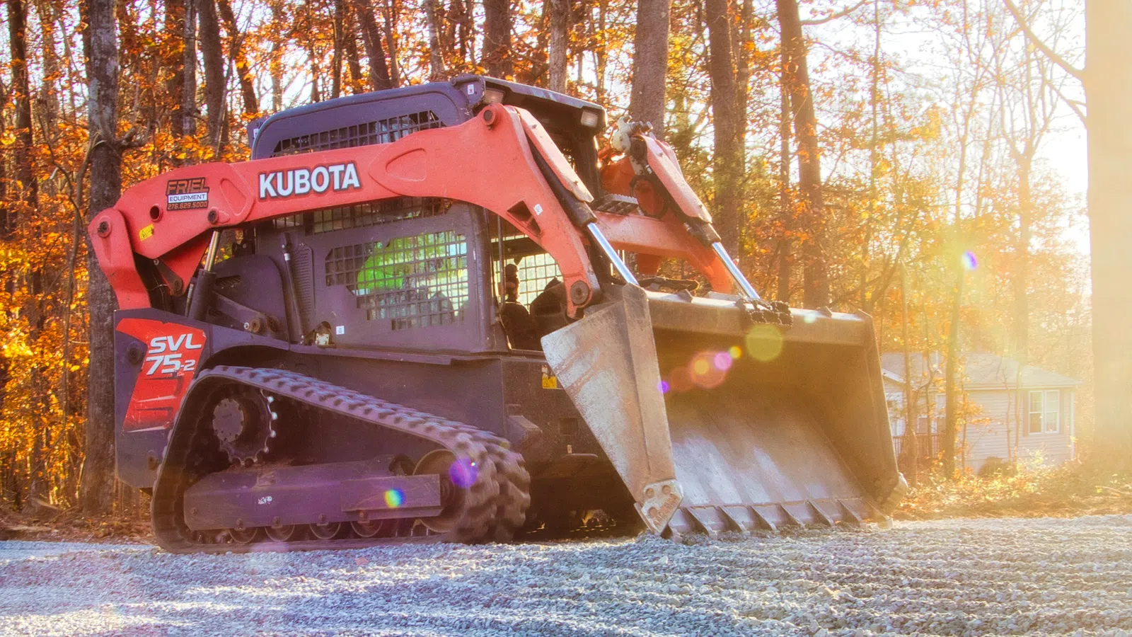 Orange Kubota skid steer loader working on gravel surface in a forested area during sunset.