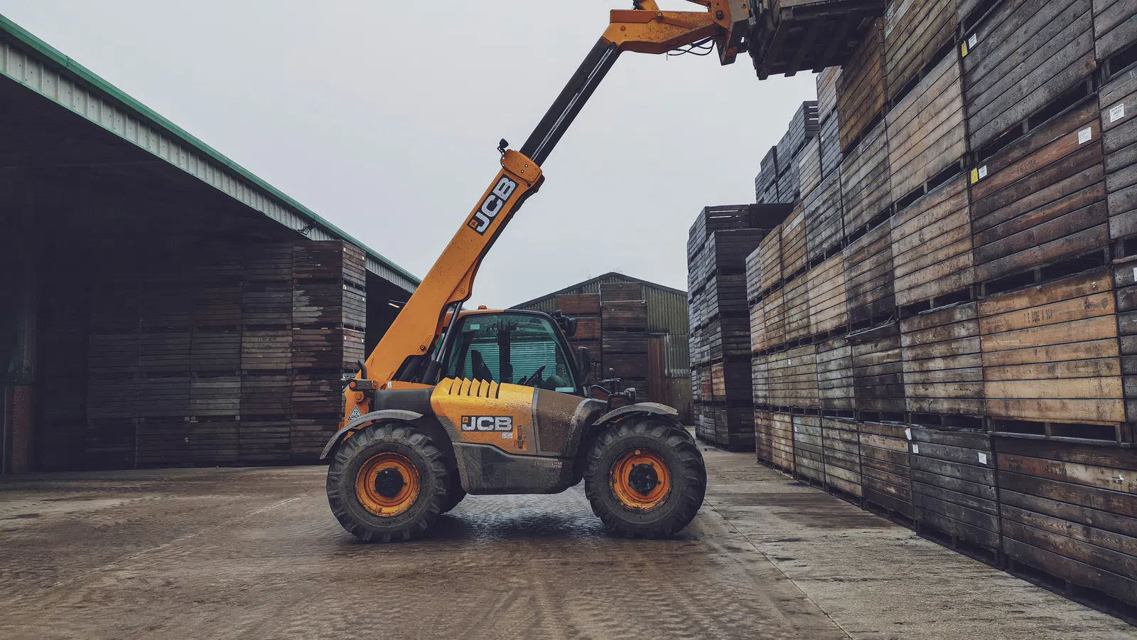 Yellow JCB forklift stacking wooden crates in an industrial warehouse yard on a cloudy day